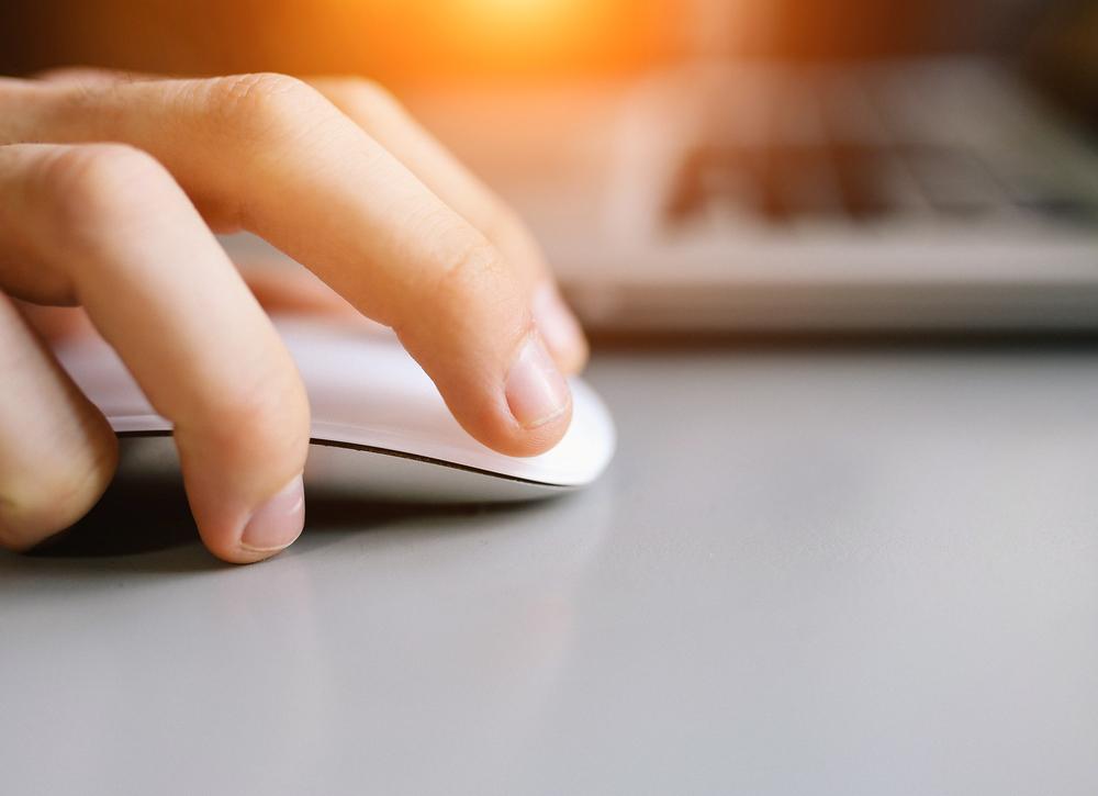 Close-up of a hand using a white computer mouse on a gray surface, with part of a laptop keyboard visible in the background and warm light in the top left corner.