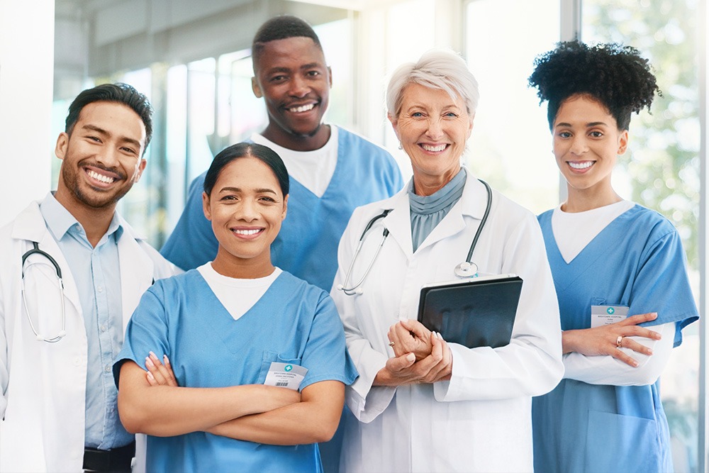 A diverse group of healthcare professionals, including doctors and nurses, smiling together in a bright medical setting. One doctor holds a tablet.